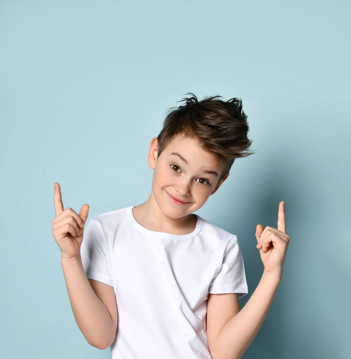 Naughty kid looking at camera with broad sly smile pointing his index fingers up getting up to mischief. Half-length portrait isolated on light blue